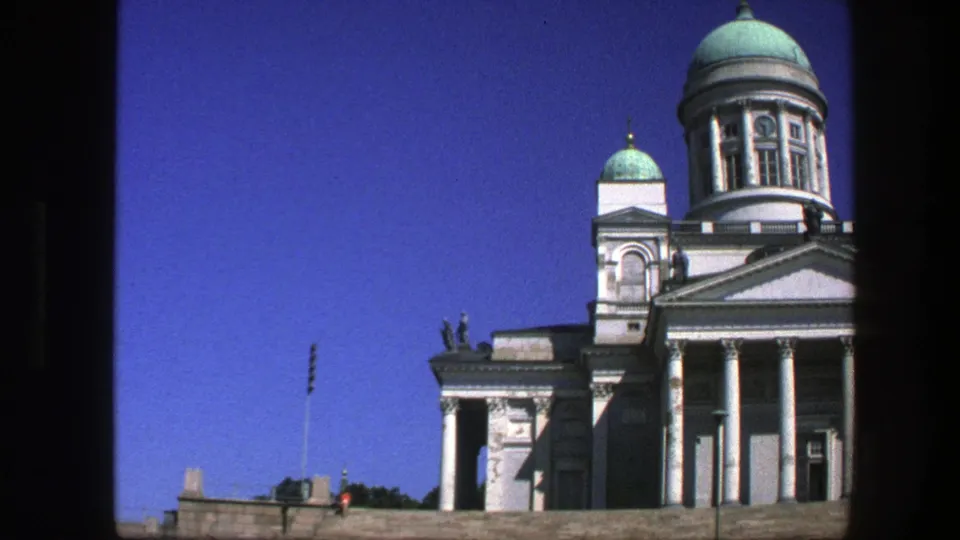 1975 8mm frame showing the side of a domed white landmark above stone steps, with tiny pedestrians near the square.