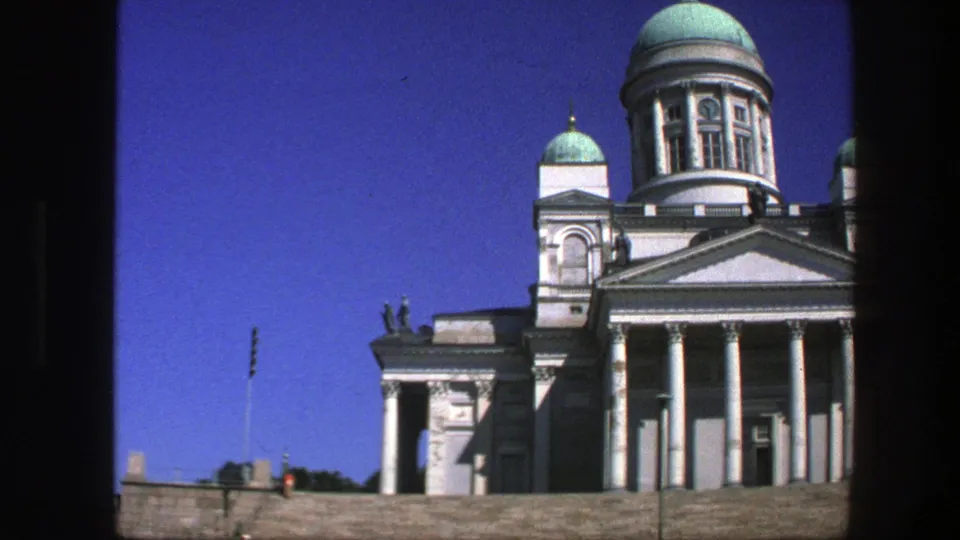 Cropped 1975 8mm view of a white domed public landmark with columns, roof statues, stone steps, and clear sky.