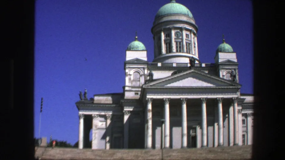 Visitor-shot 8mm frame of Helsinki Cathedral, showing the columned front, green domes, blue sky, and distant people on the plaza.