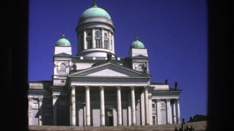1970s 8mm frame of Helsinki Cathedral with green domes, white facade, columned entrance, and distant visitors near the edge.