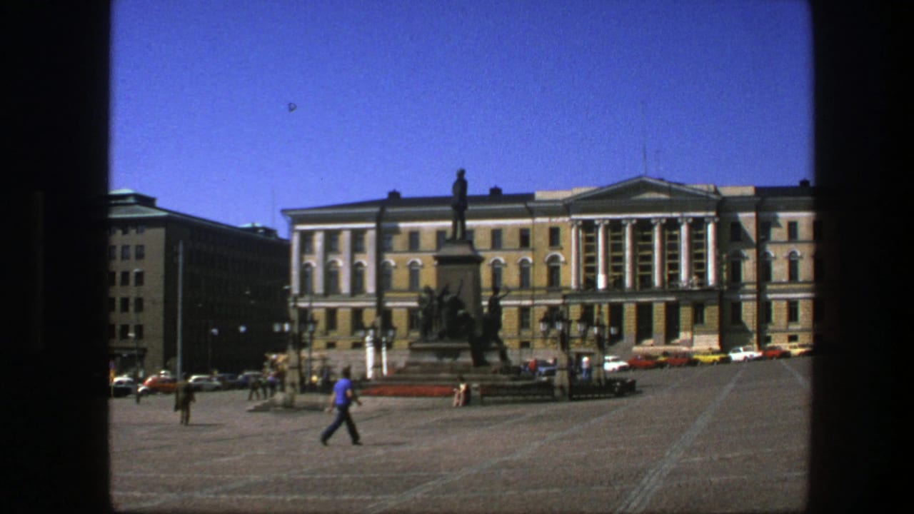 1975 Sweden home-movie frame showing a broad square, people walking, a tall monument, and a formal building under blue sky.