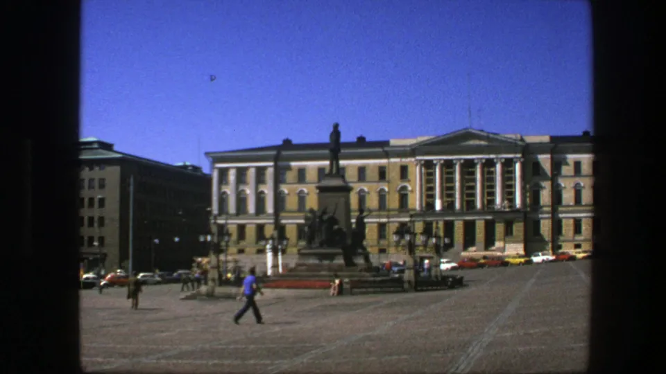 1975 Sweden home-movie frame showing a broad square, people walking, a tall monument, and a formal building under blue sky.