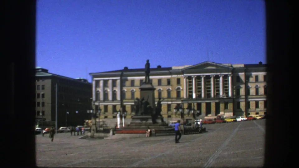 1975 Sweden home-movie still showing a sunlit square, distant pedestrians, a statue monument, and a large institutional facade.