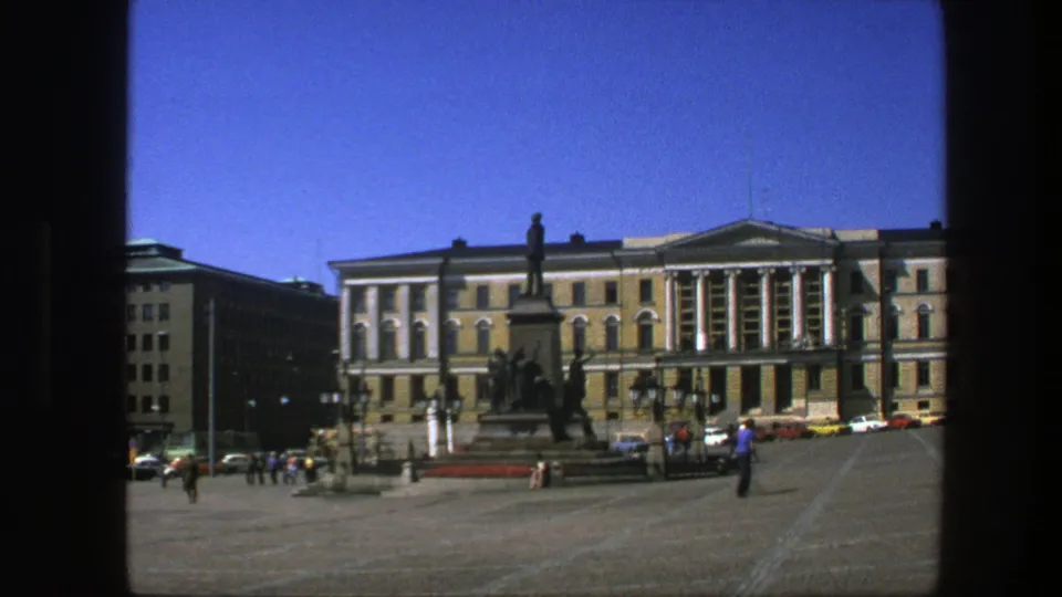 1975 Sweden frame showing pedestrians crossing a public square before a tall statue monument and a classical building.