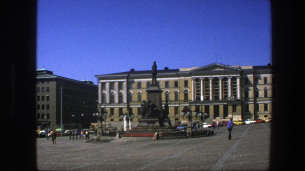 Paved city square with a monument, cars, pedestrians, and a large classical building with columns.