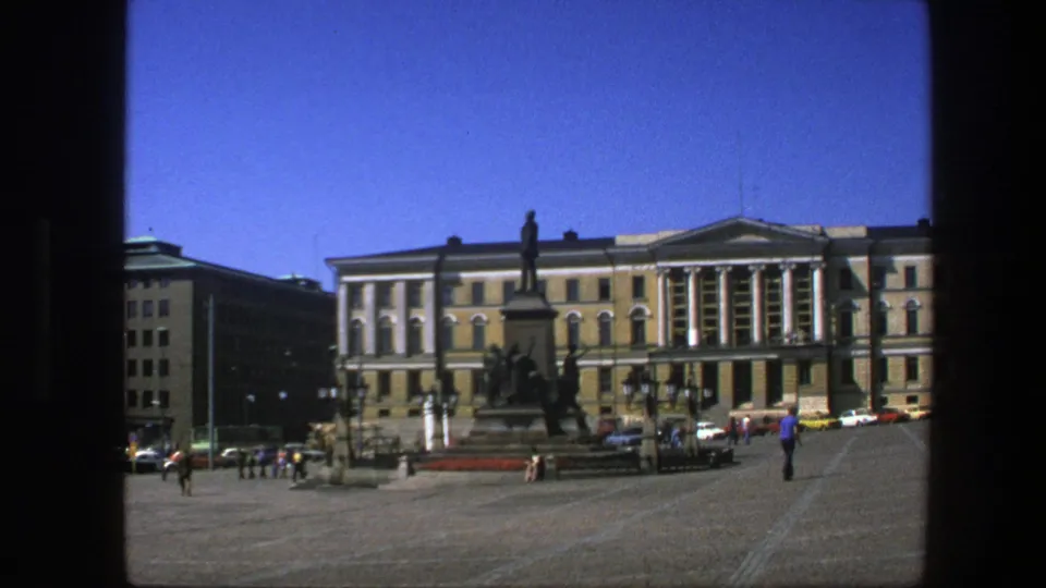 1975 Sweden home-movie frame showing a civic square, central monument, pedestrians, cars, and a large columned building in sunlight.