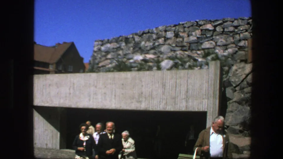 1975 Sweden frame with several adults walking out of a modern concrete tunnel set against older stonework.