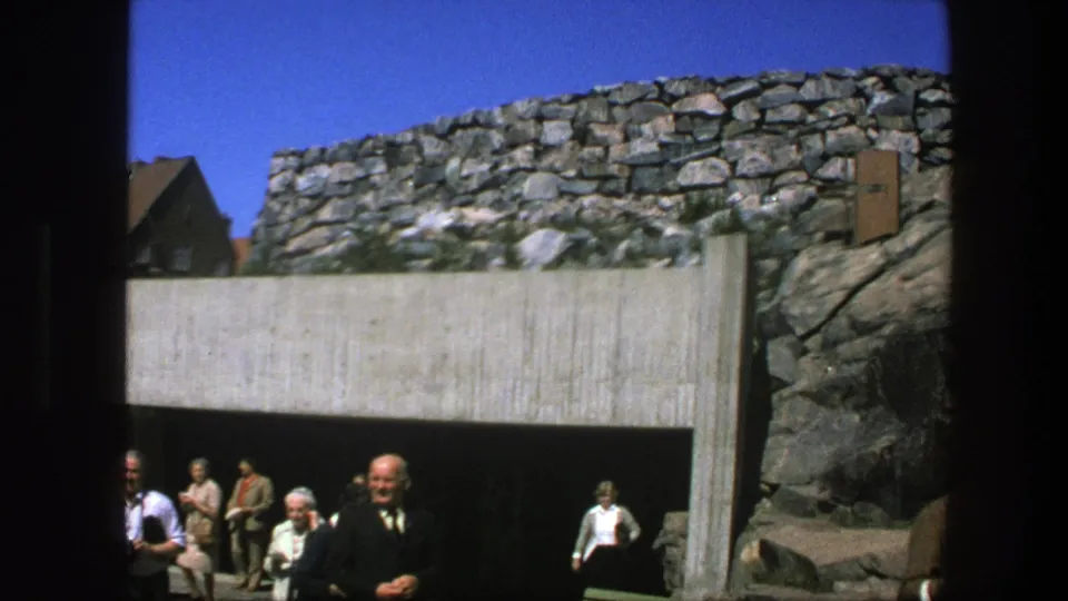 1975 Sweden home-movie frame showing a tunnel entrance below stonework, with adults gathered and walking nearby.