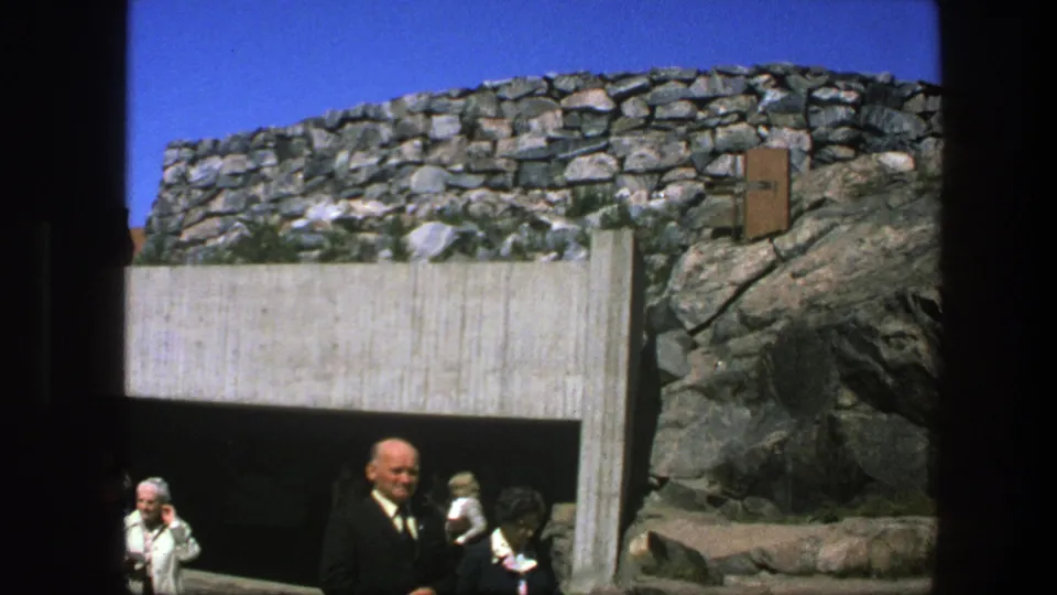 1975 Sweden frame with older adults near a tunnel opening, rough stonework, exposed rock, and bright blue sky.