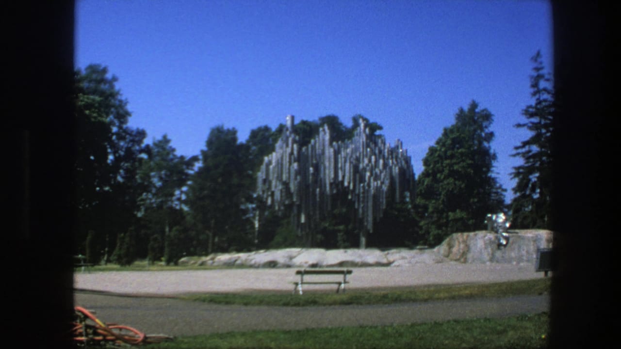 Park view with metal pipe sculpture, trees, blue sky, paved ground, and a wooden bench or picnic table.