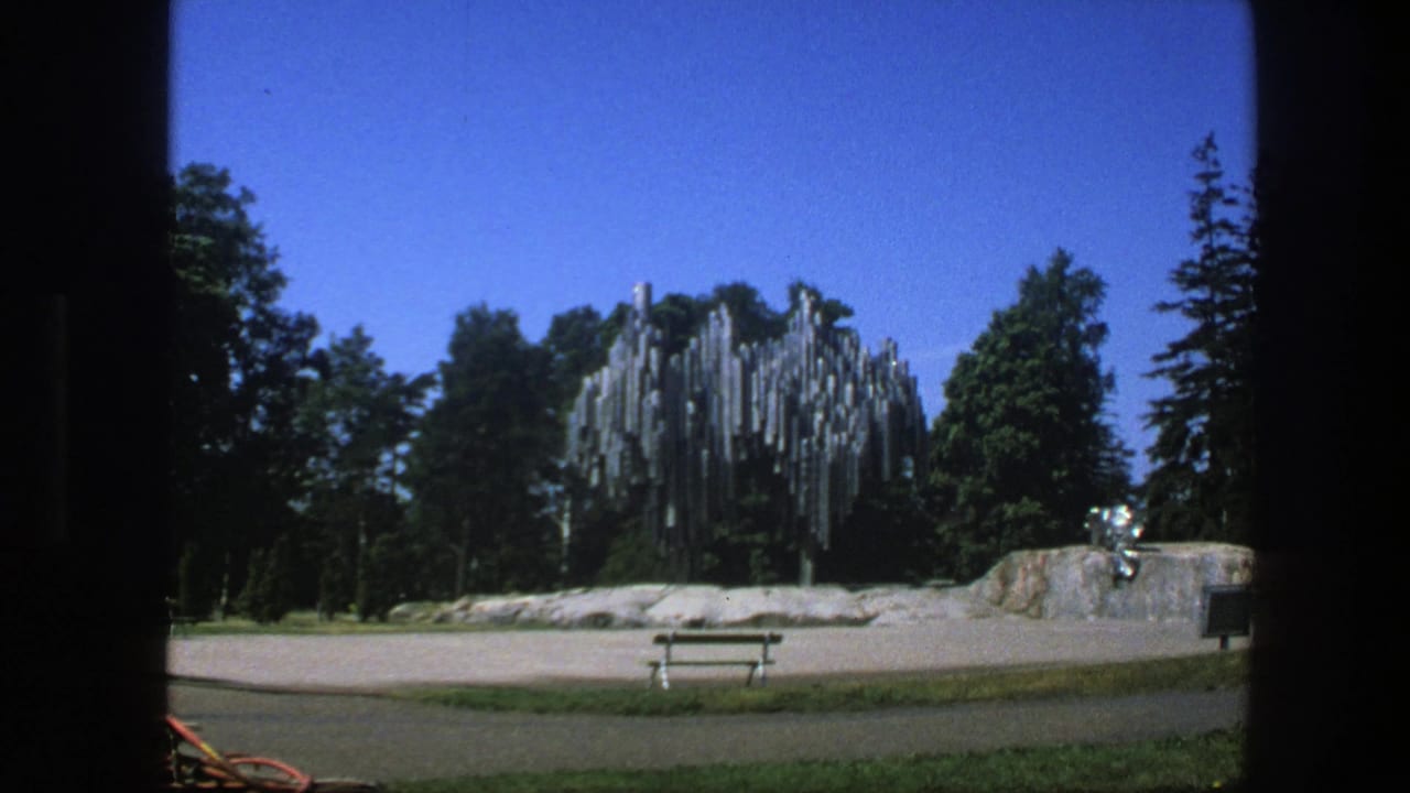Metal sculpture in a park with trees, a bench or table, paved ground, grass, and clear sky.