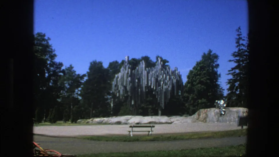 1975 Sweden park view on 8mm film, with a vertical metal sculpture against trees and a small seating area in front.