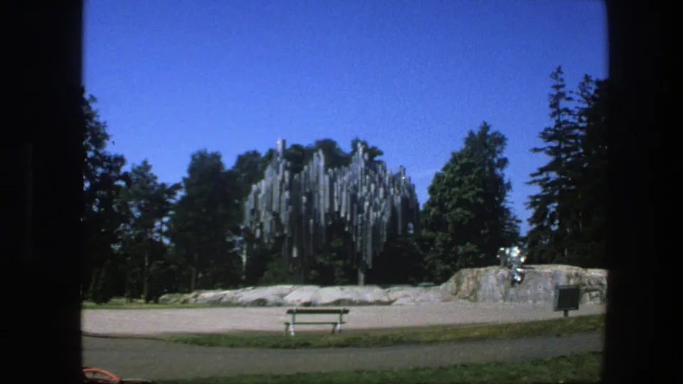 1975 Sweden home-movie frame with a modern metal sculpture in a green park, seen from a steady distant viewpoint.