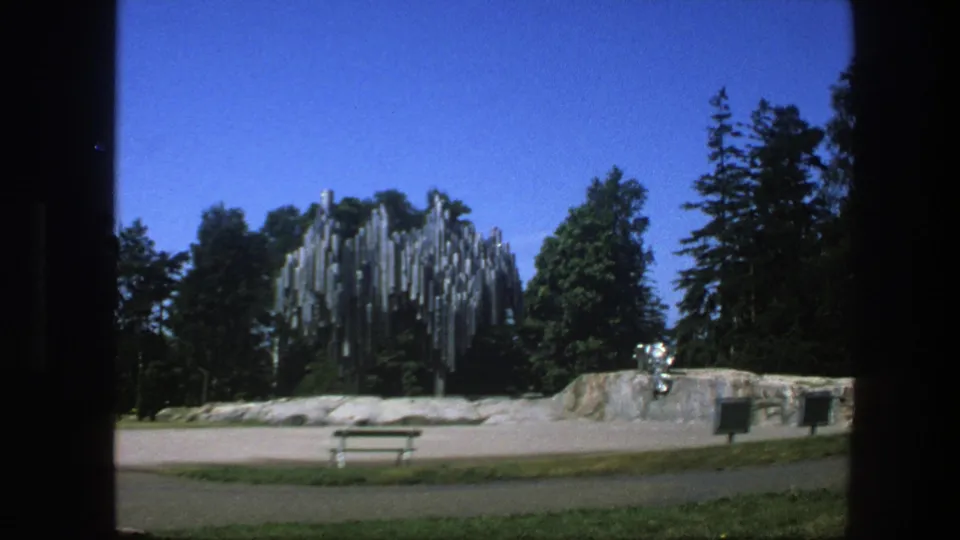 1975 Sweden 8mm frame of a quiet public park, with a metal pipe sculpture, trees, seating, grass, and paved foreground.
