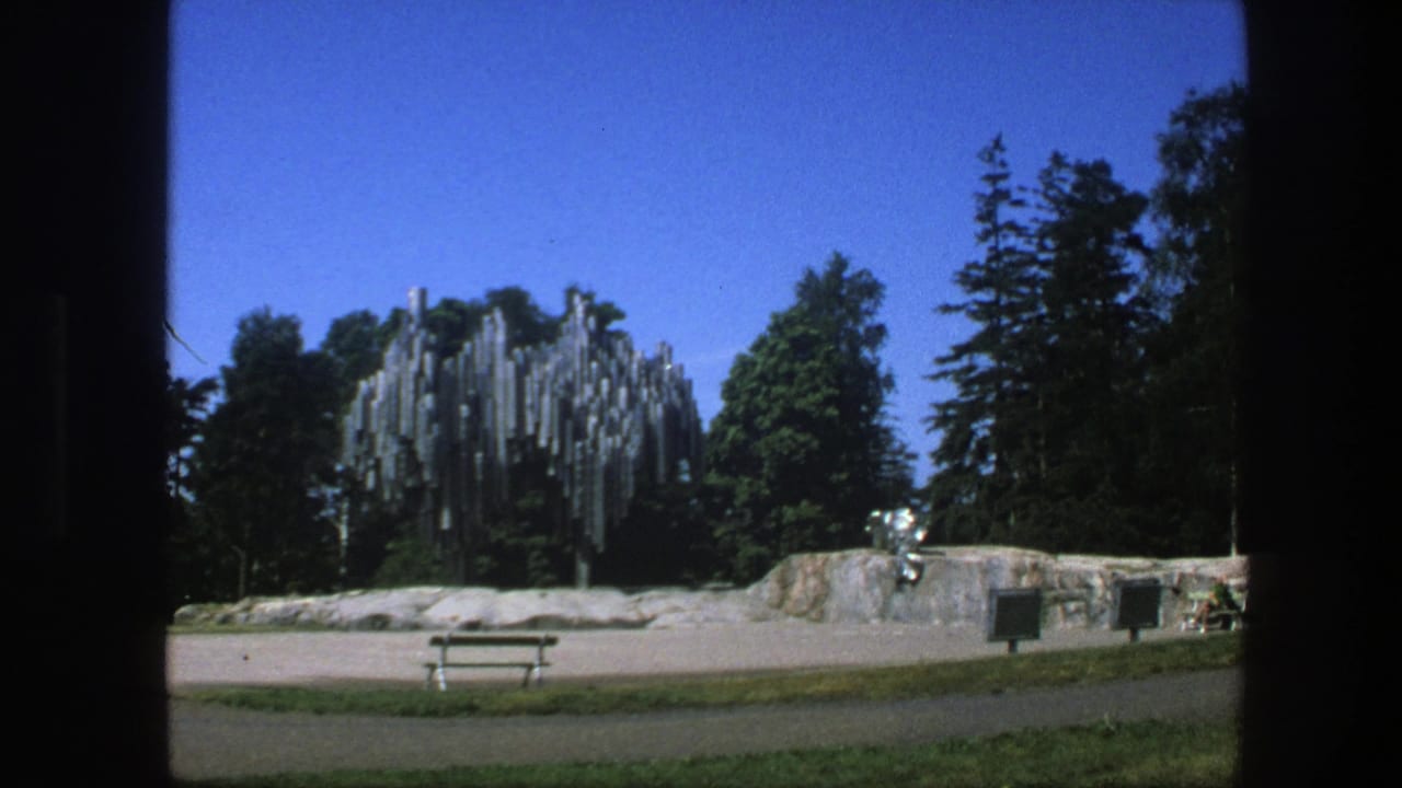 Park scene with reflective metal sculpture, stone wall, bench, trees, and blue sky.