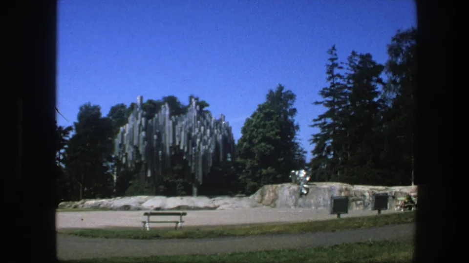 1975 Swedish 8mm frame showing a park with a reflective tube-like sculpture, low stone wall, bench, trees, and clear blue sky.