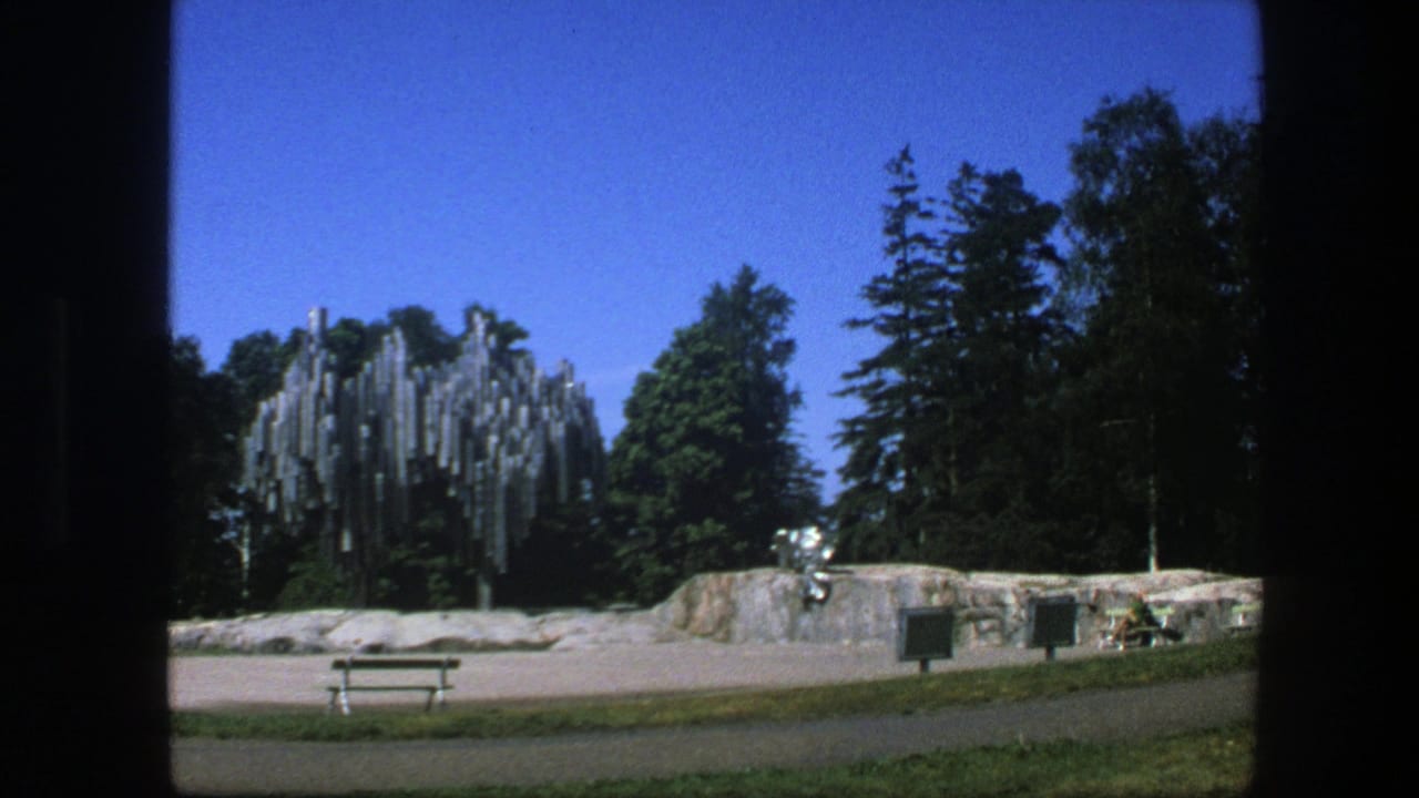 1975 Swedish park view with a modern reflective sculpture beside a low rock wall, with trees, bench, path, and blue sky visible.