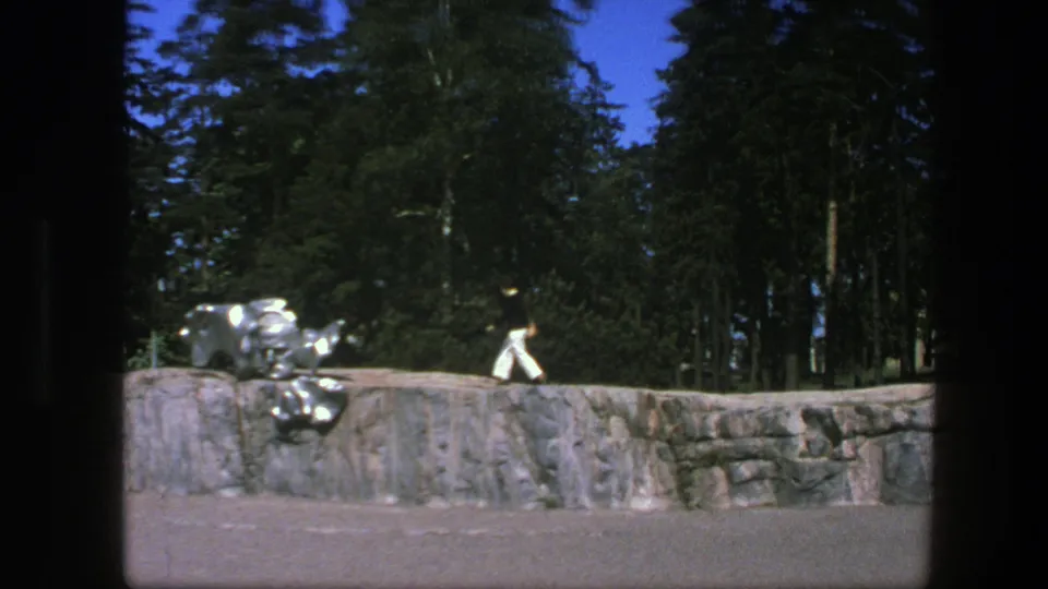 1975 Swedish 8mm frame with a distant person in white trousers walking along a low rock wall near reflective sculpture elements and tall trees.