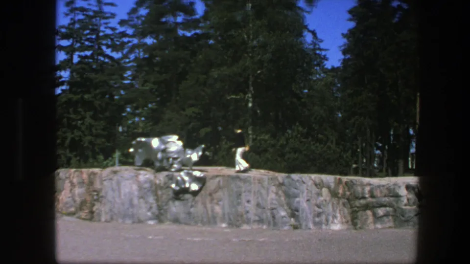 1975 Swedish park footage frame showing a distant person walking beside a low stone wall, with reflective sculpture pieces and dense trees behind.