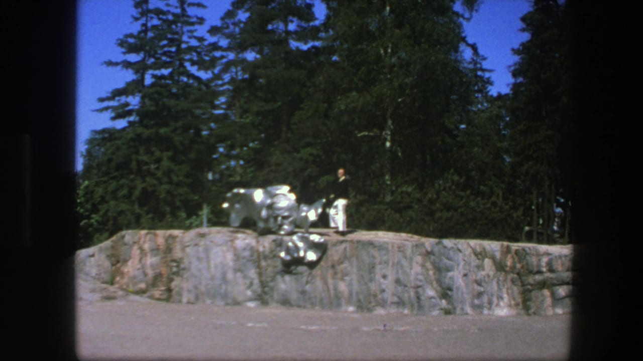 1975 Sweden 8mm frame showing a distant person in white pants beside a reflective silver sculpture on a rocky platform under trees.