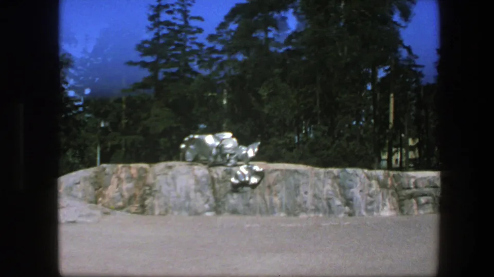 1970s 8mm view of a silver abstract sculpture set on a rocky outdoor platform with dense trees and bright blue sky.