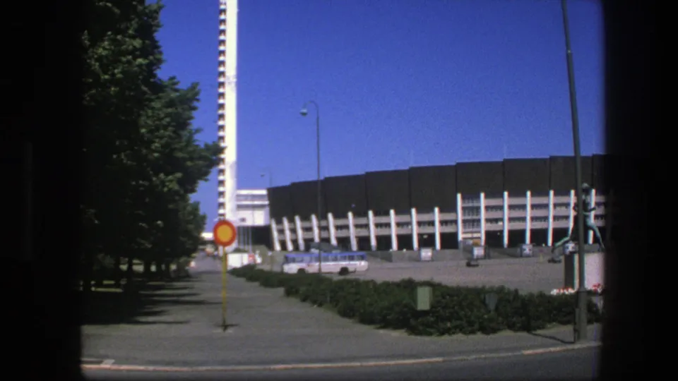 1975 Sweden 8mm frame showing a modern street-side building, sidewalk, trees, traffic sign, parked cars, and clear blue sky.