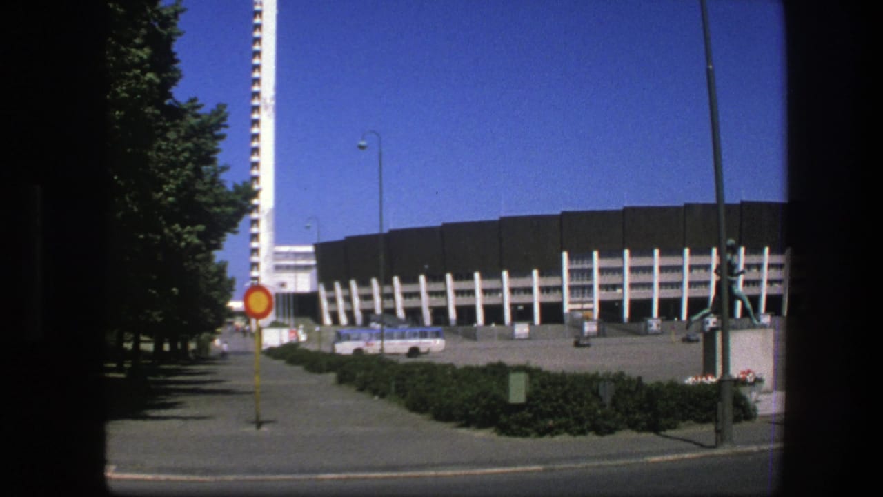 Stadium exterior with white supports, tall tower, trees, road, sign, and statue in front.
