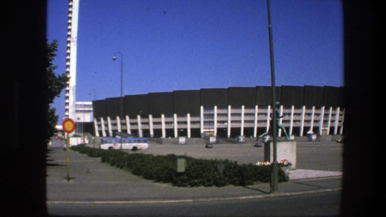 Wide stadium front with tower, statue, sign, hedges, paved plaza, and parked bus.