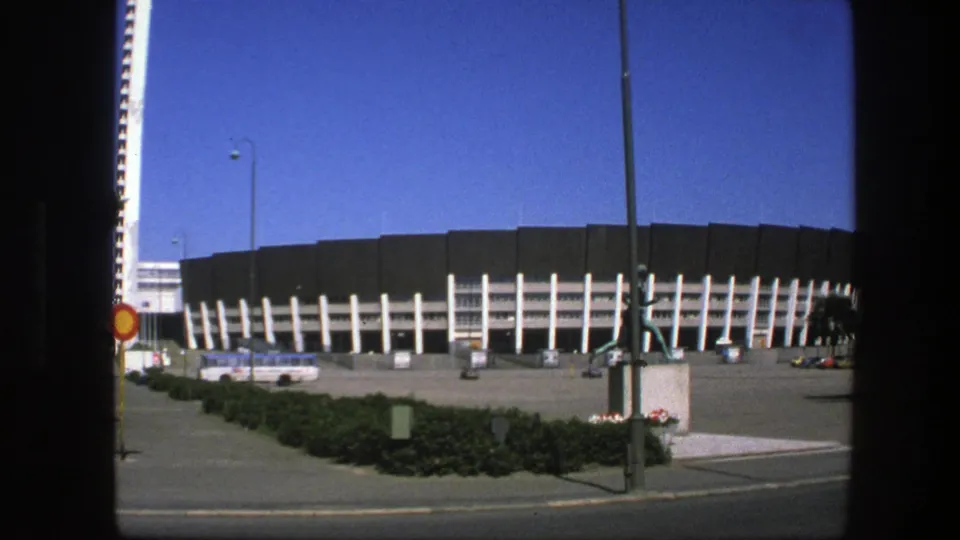 1975 Sweden 8mm still showing the stadium facade from the street, with white supports, tower edge, runner statue, bus, sign, and planted median.