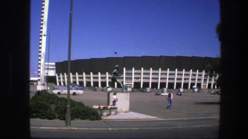 1975 Sweden frame of the stadium frontage with the runner statue in the foreground, bus by the facade, tower, streetlight, and distant pedestrians.