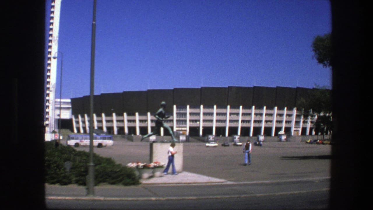 Stadium plaza with running athlete statue, pedestrians, bus, cars, and clear blue sky.