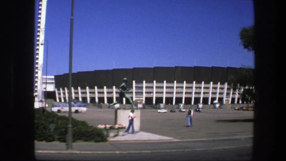 1975 Sweden stadium exterior with a running athlete statue in front, pedestrians crossing the plaza, a bus, cars, and a tall white tower.