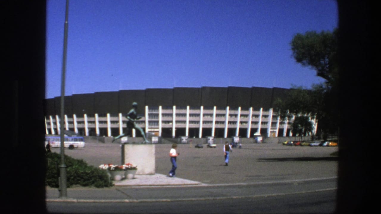 1975 Sweden view of a stadium plaza with a bronze running athlete statue, scattered pedestrians, parked cars, and a bus in bright sun.