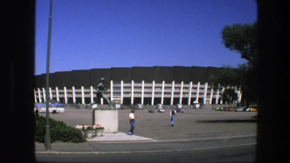 1975 Sweden view of a stadium plaza with a bronze running athlete statue, scattered pedestrians, parked cars, and a bus in bright sun.