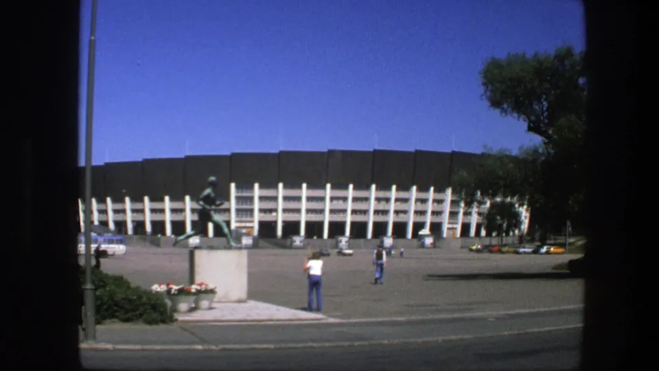 1975 Sweden stadium exterior showing the running athlete statue, a few people in the plaza, flower planters, road edge, and strong midday light.