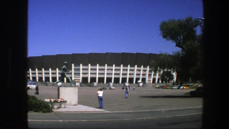 1975 Sweden stadium plaza with the running athlete statue in front, distant pedestrians, trees at the edge, parked cars, and a bright blue sky.