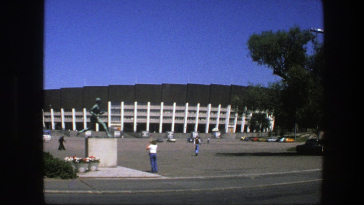 Stadium exterior with white columns, runner statue, pedestrians, cars, and trees across a street.