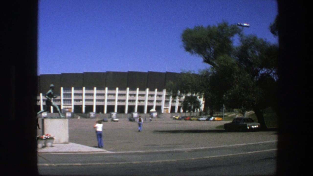 Wide view of stadium facade, statue, plaza, pedestrians, parked cars, and roadside trees.