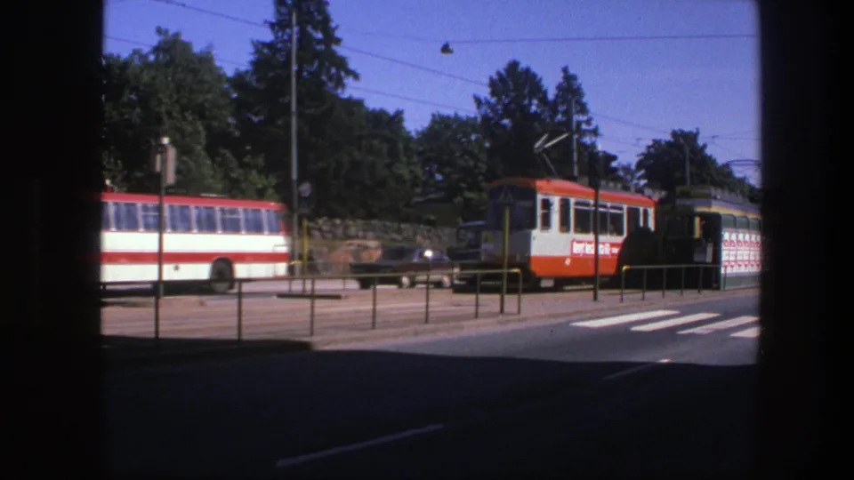 1975 Sweden 8mm street view showing a red-and-white trolley, a matching bus, overhead wires, roadside railings, trees, and cars.