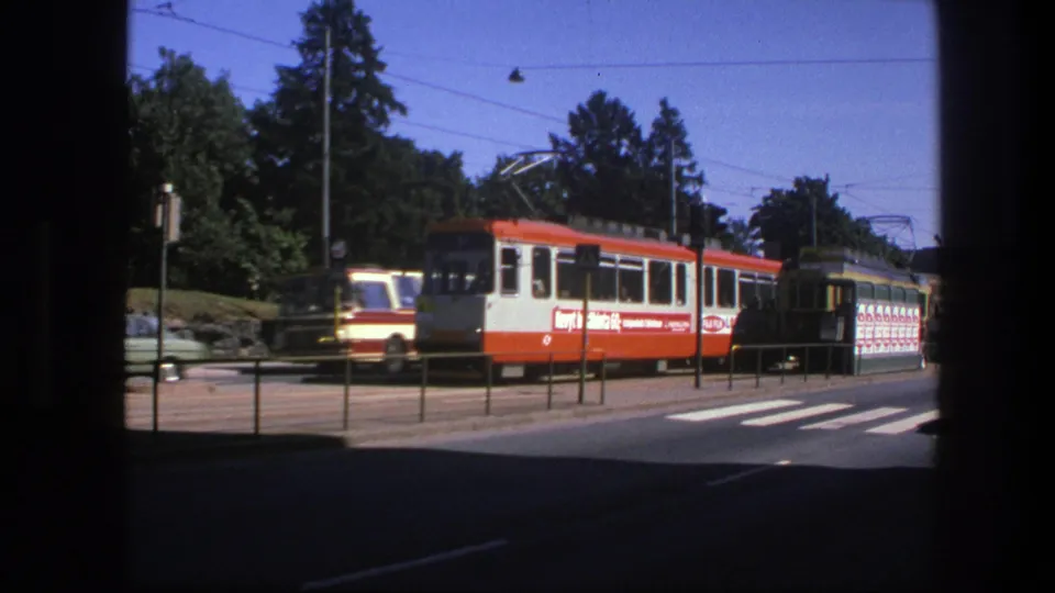1975 Sweden frame showing a red-and-white trolley beside a similar bus, with classic cars, overhead wires, trees, street railings, and a crosswalk.