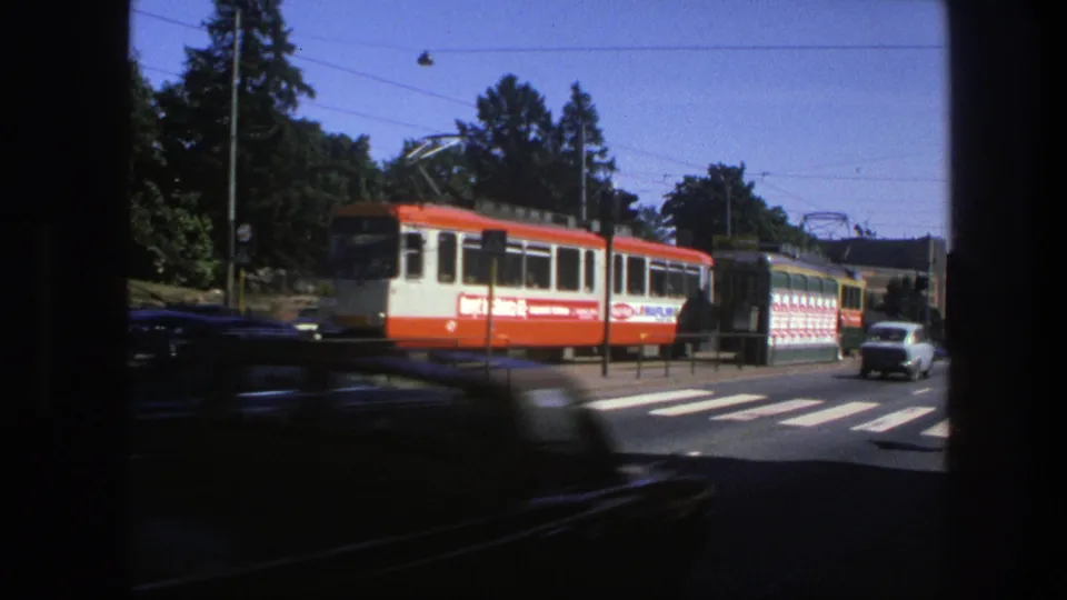1975 Sweden tram stop scene with an orange-and-white tram, bus, overhead wires, and a blurred car crossing the frame.