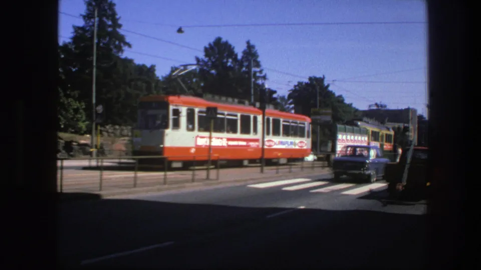 1975 Sweden street footage showing an electric tram, crosswalk markings, roadside railings, trees, and a dark car in the lane.