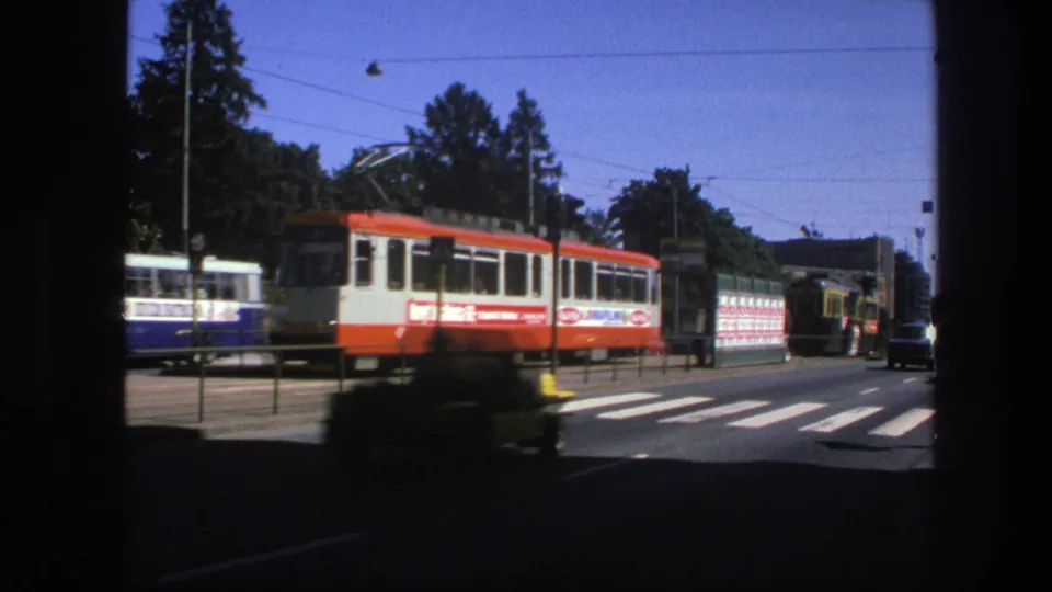 1975 Sweden street scene with an electric tram, another transit vehicle, overhead wires, crosswalk, trees, and passing traffic.