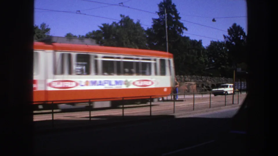 1975 Sweden 8mm frame showing the tram passing close to camera, with trees, railings, a white car, and dark frame borders.