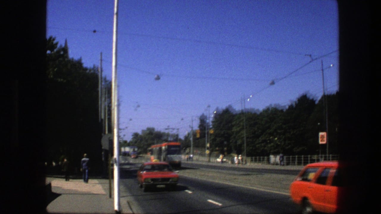 1975 Sweden street view with a red car, distant red-and-white trolley, overhead wires, trees, sidewalk, and pedestrians.