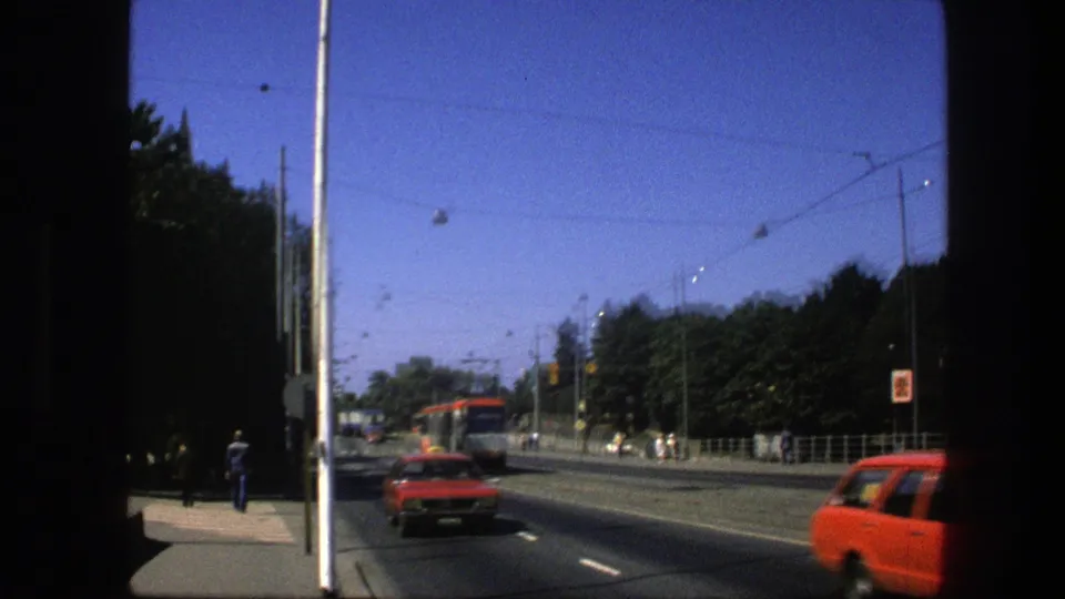 1975 Sweden street view with a red car, distant red-and-white trolley, overhead wires, trees, sidewalk, and pedestrians.