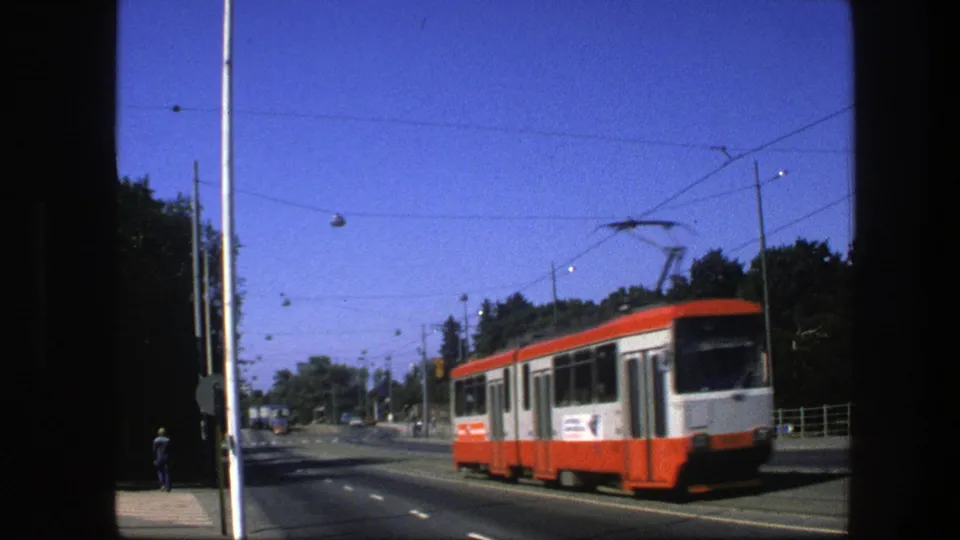Red-and-white trolley car passes near the camera on a 1975 Sweden city avenue with wires, streetlamps, trees, and sidewalk pedestrians.