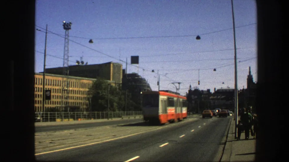 1975 Sweden city avenue view with the red-and-white trolley moving away beside buildings, overhead wires, streetlamps, and pedestrians.