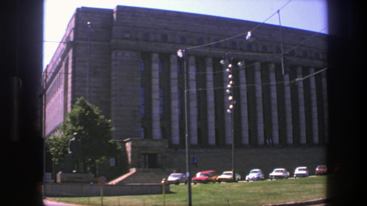 Large stone public hall with columns, parked cars, lawn, statue, and overhead street wires.