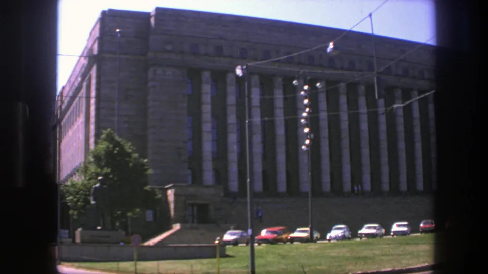 1975 Sweden 8mm frame showing a monumental stone public hall, vertical columns, a row of cars, lawn, statue, and overhead wires.